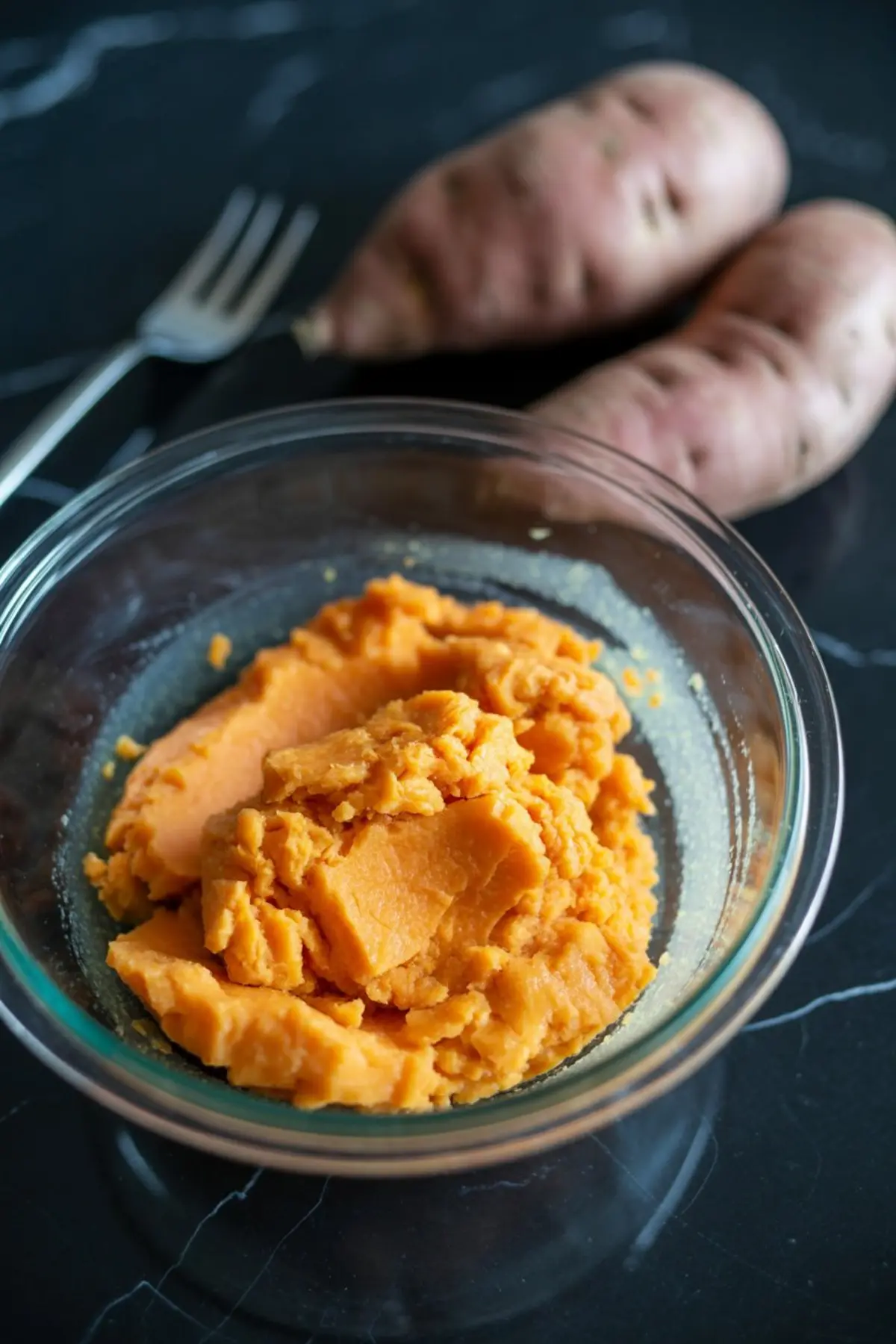 Glass bowl filled with mashed orange sweet potatoes placed on a black marble surface, with raw sweet potatoes and a fork in the background for a rustic cooking prep scene.