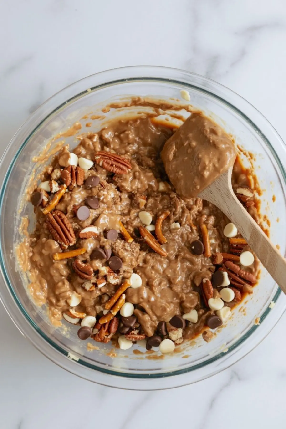 Mixing bowl filled with Texas Trash Pie batter, featuring a sticky blend of crushed pretzels, pecans, white chocolate chips, and milk chocolate chips stirred with a wooden spatula.