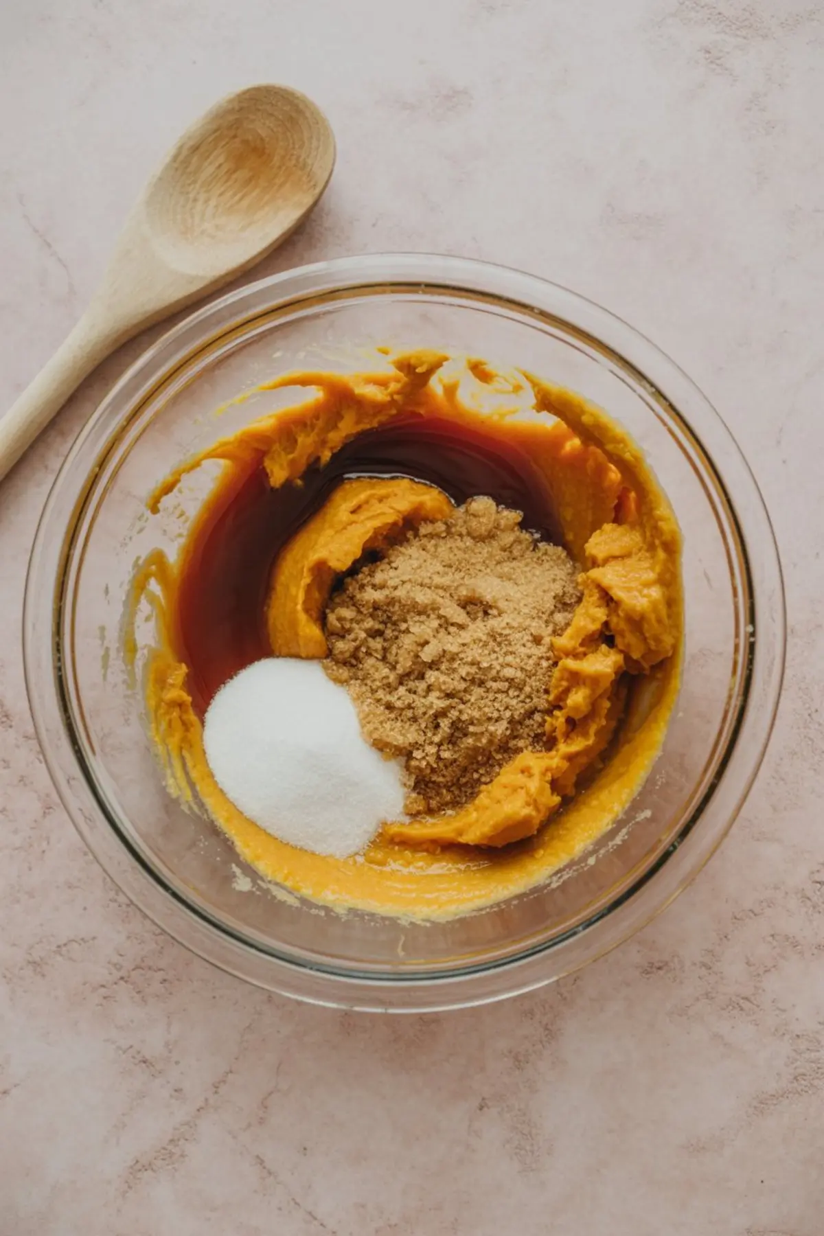 Glass mixing bowl with pumpkin purée, brown sugar, white sugar, and vanilla extract set on a pale marble background with a wooden spoon nearby.