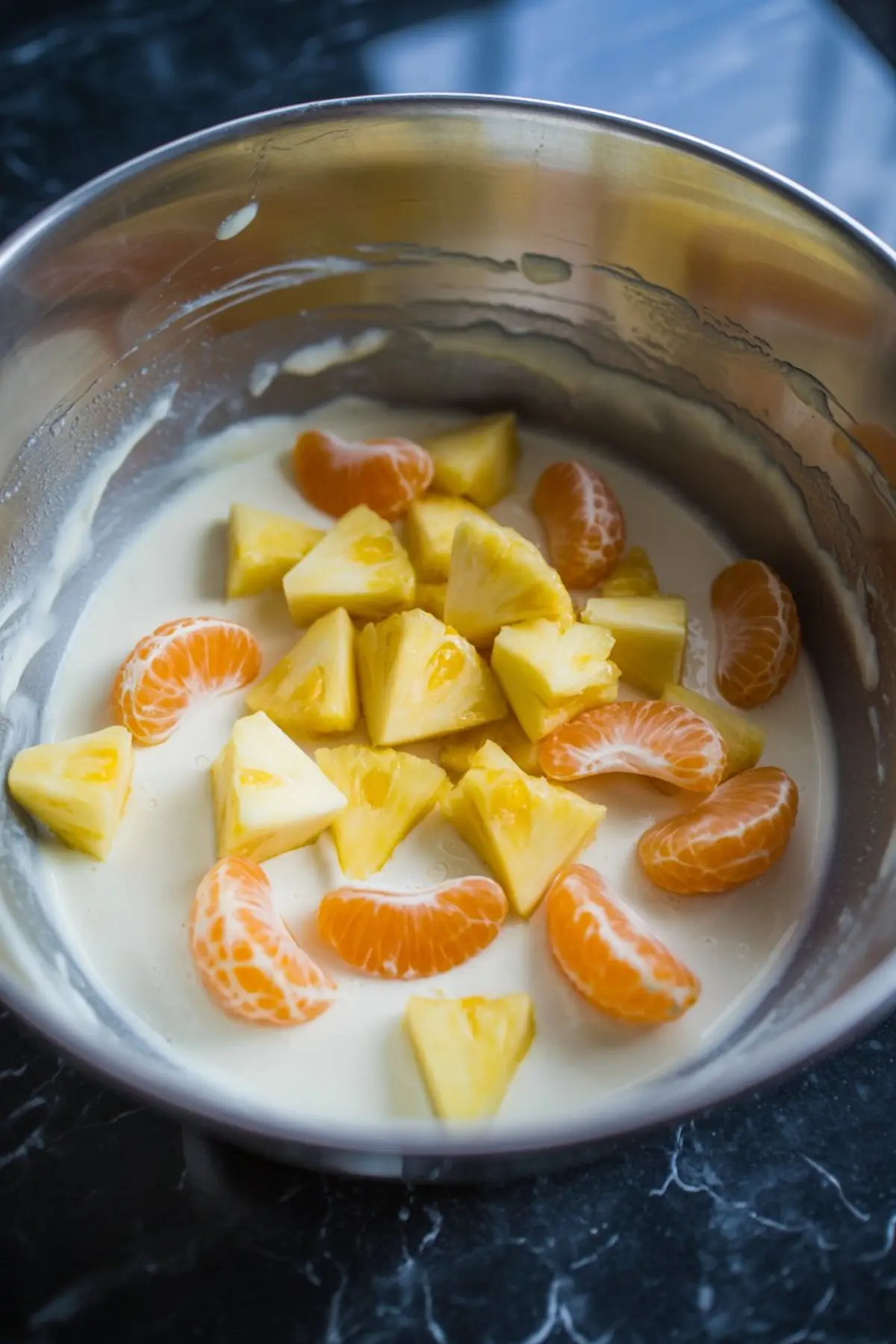 Metal mixing bowl filled with pineapple chunks and mandarin orange slices in a creamy base, ready to be mixed into a fruit salad.