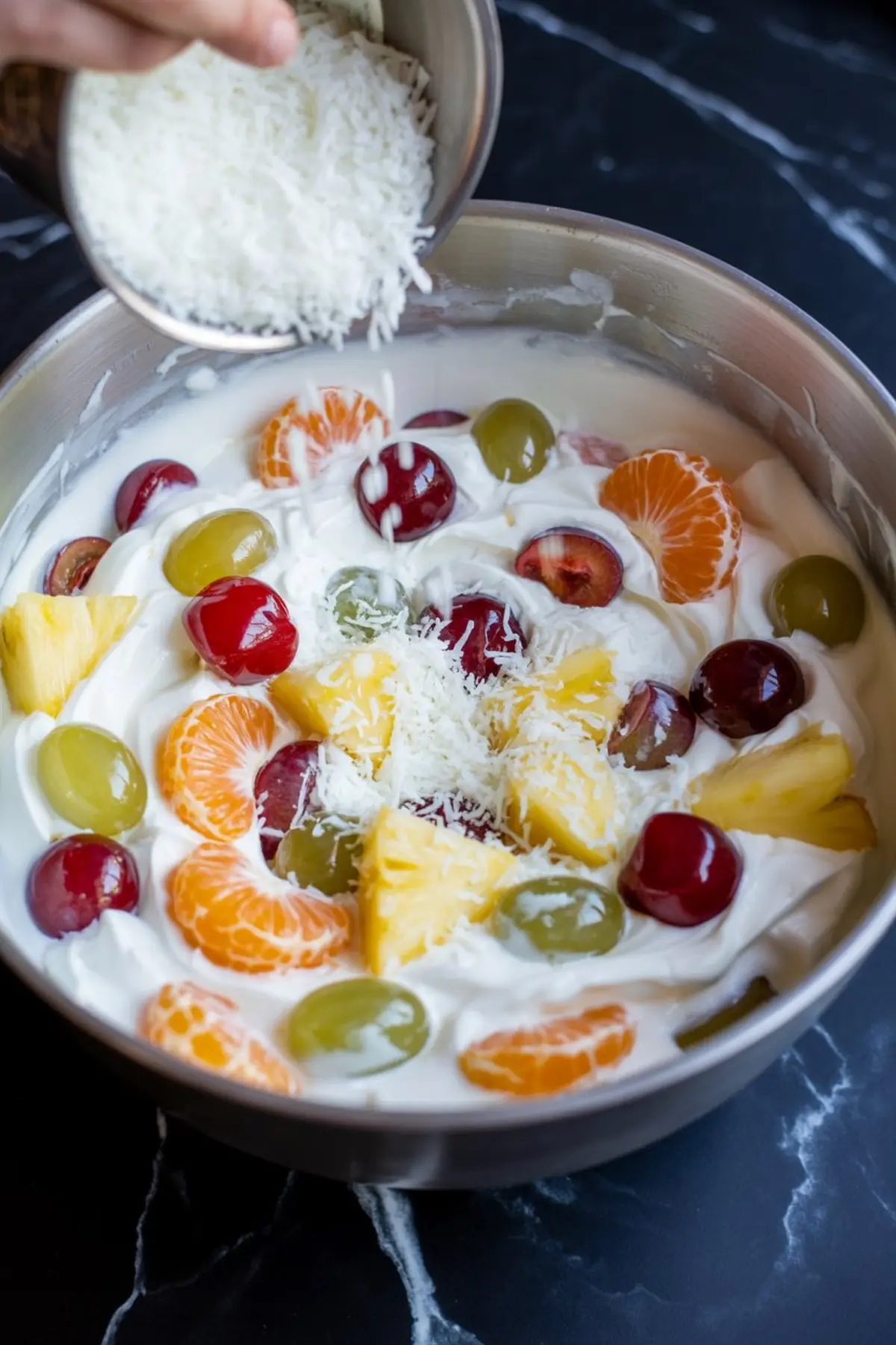 Large bowl of creamy fruit salad topped with pineapple, grapes, mandarins, cherries, and a sprinkle of shredded coconut being added.