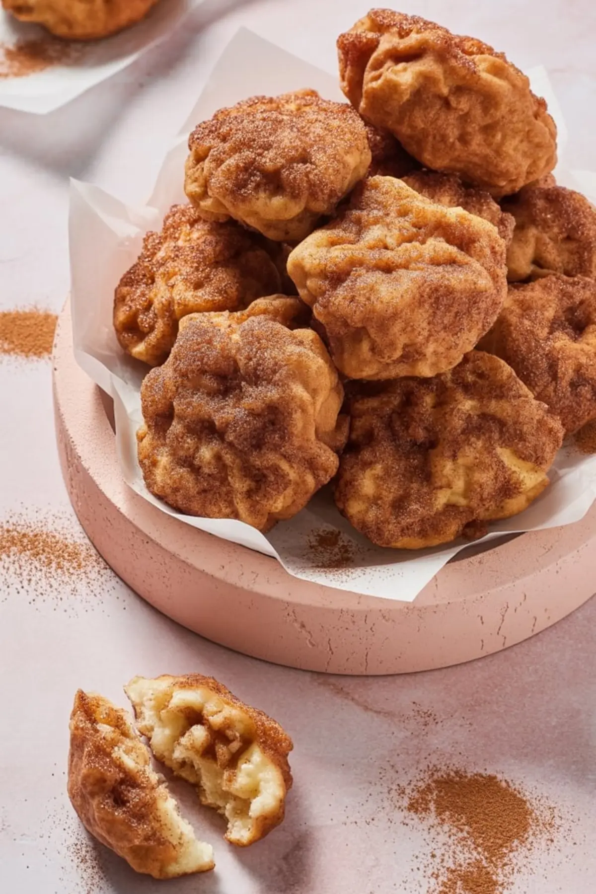 Stack of freshly fried apple fritters covered in cinnamon sugar, placed in a parchment-lined bowl with one fritter broken open to show the tender apple-filled interior.