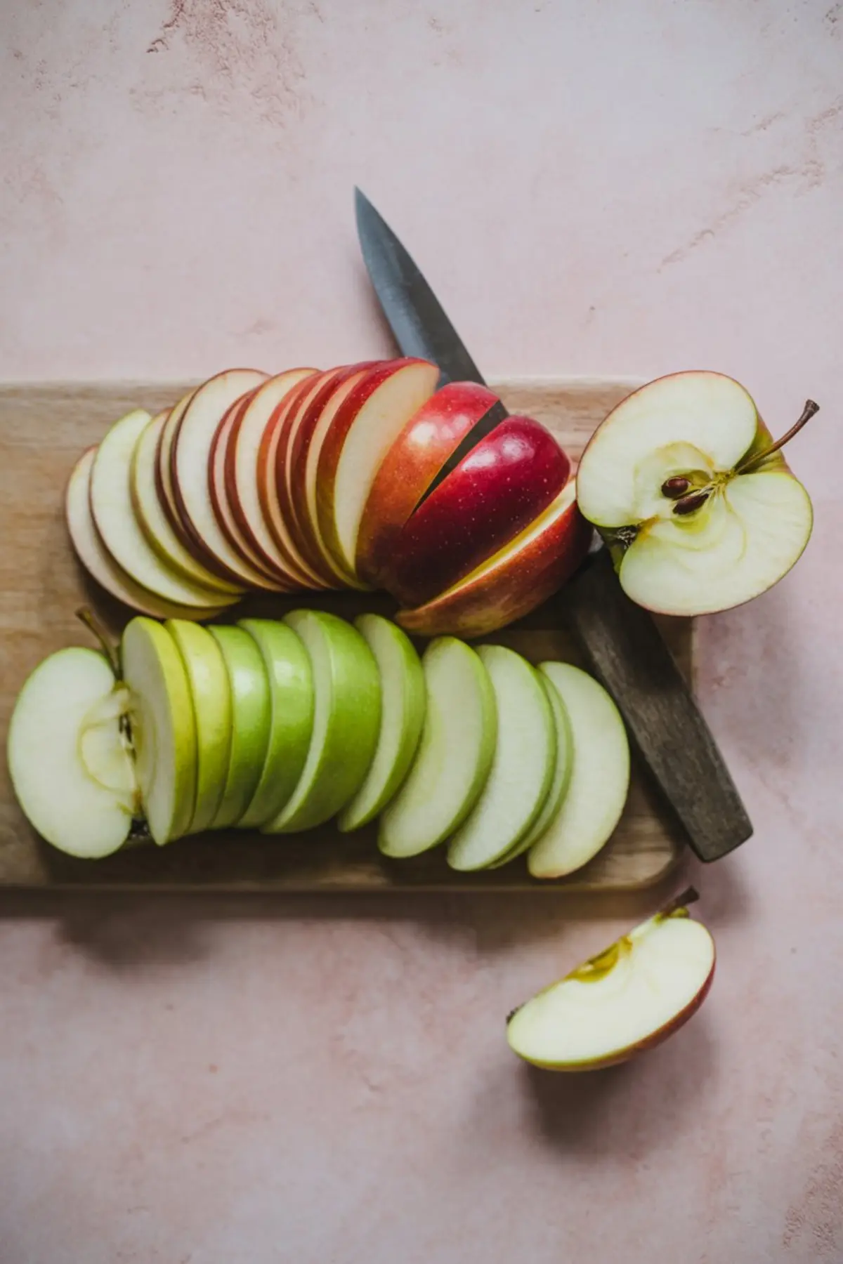 Wooden cutting board with neatly sliced red and green apples next to a halved apple and a paring knife, all set on a light pink background.