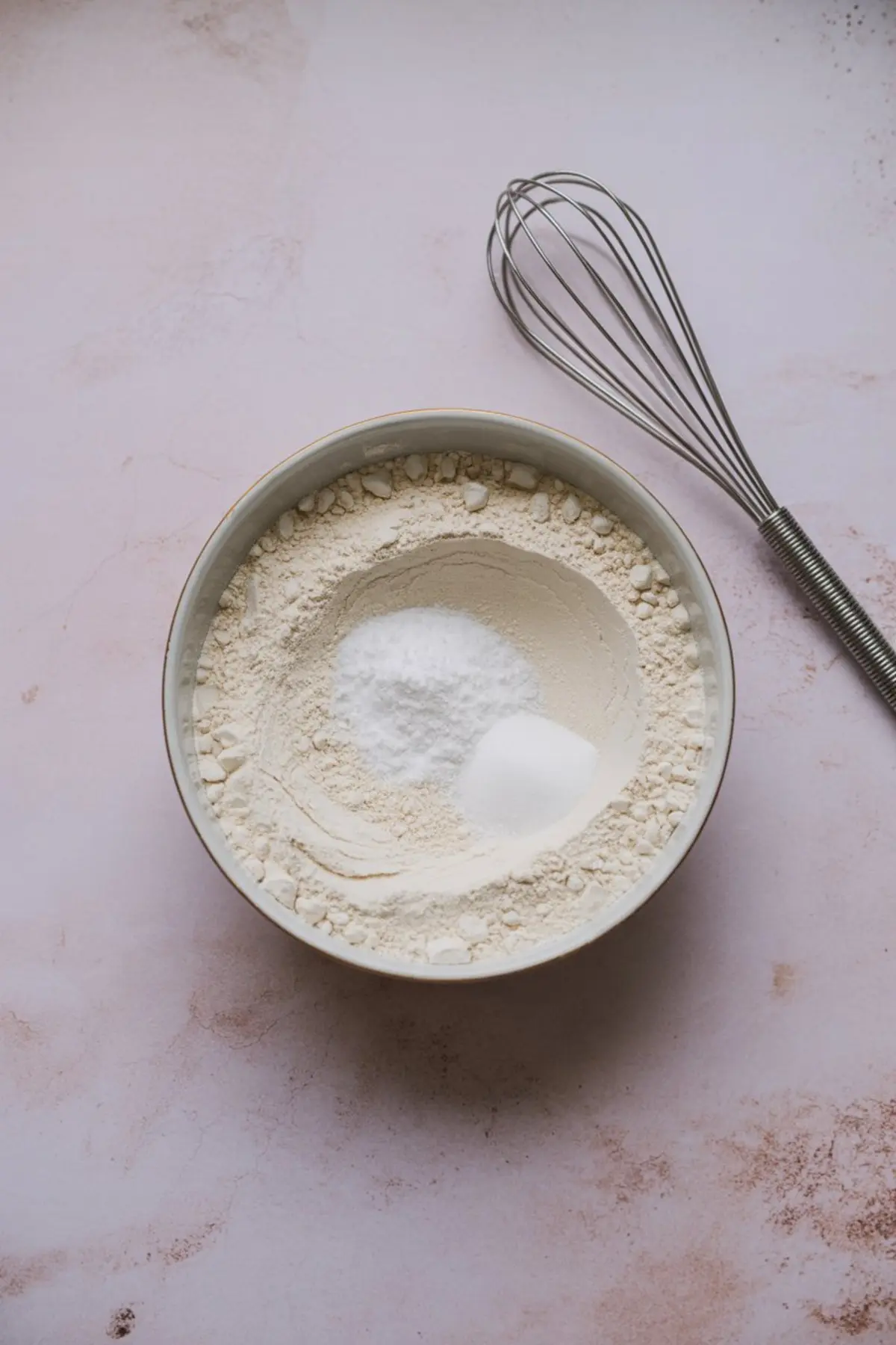 Mixing bowl filled with flour, baking powder, and salt, placed on a pink surface beside a metal whisk, prepared for butterscotch cookie recipe.
