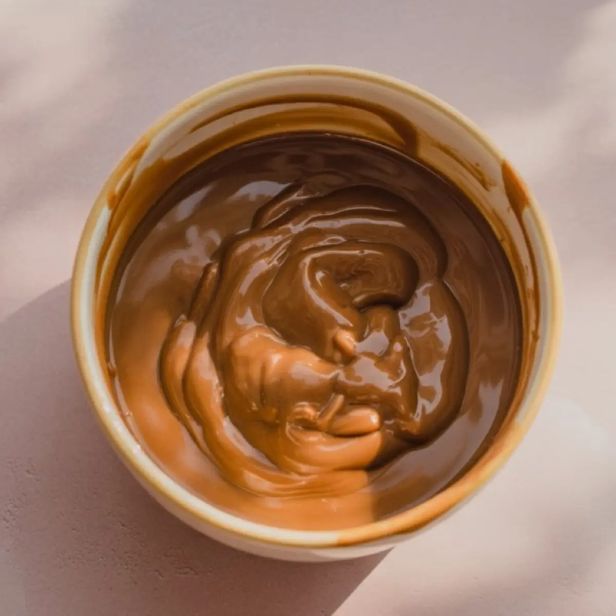 Bowl of melted butterscotch chips swirled into a smooth, glossy texture, placed on a pale pink background with soft shadows.
