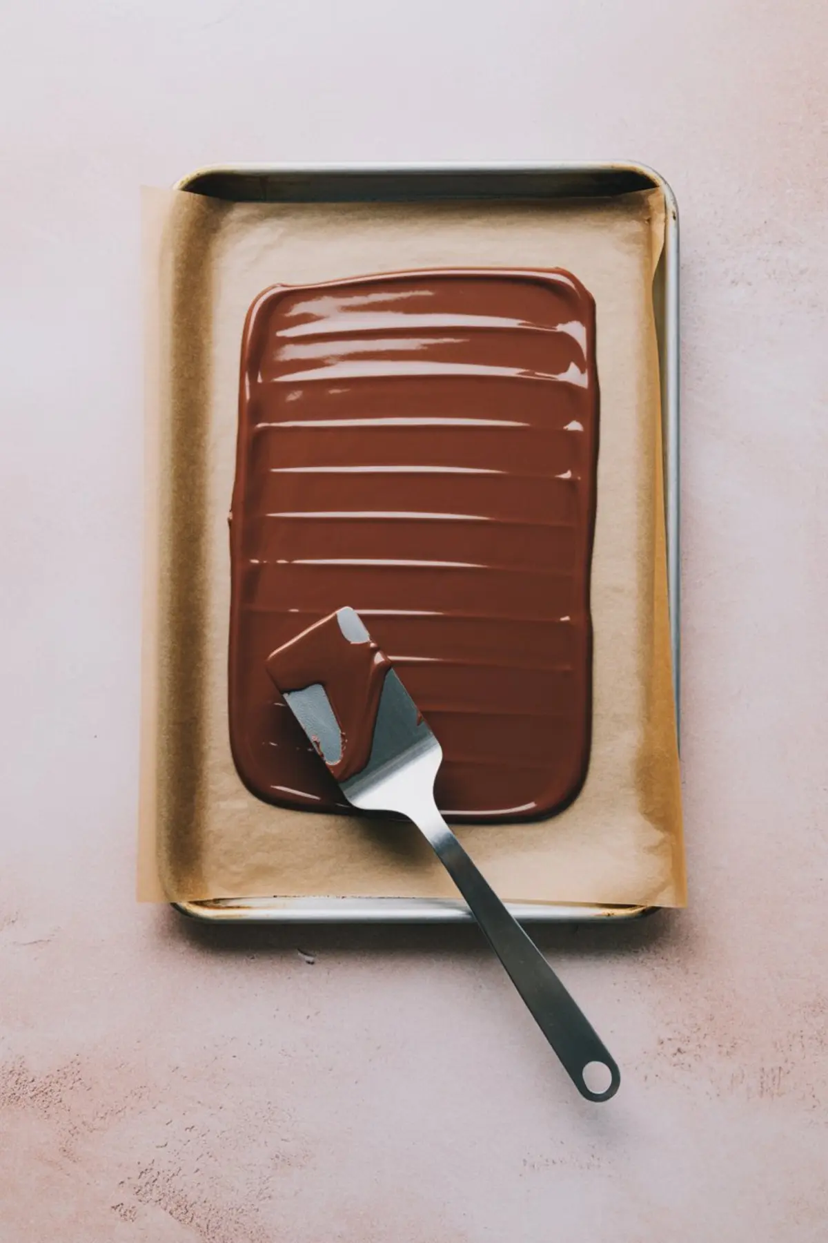 Sheet pan lined with parchment paper holding freshly spread melted chocolate, smoothed with a metal spatula for making chocolate bark.
