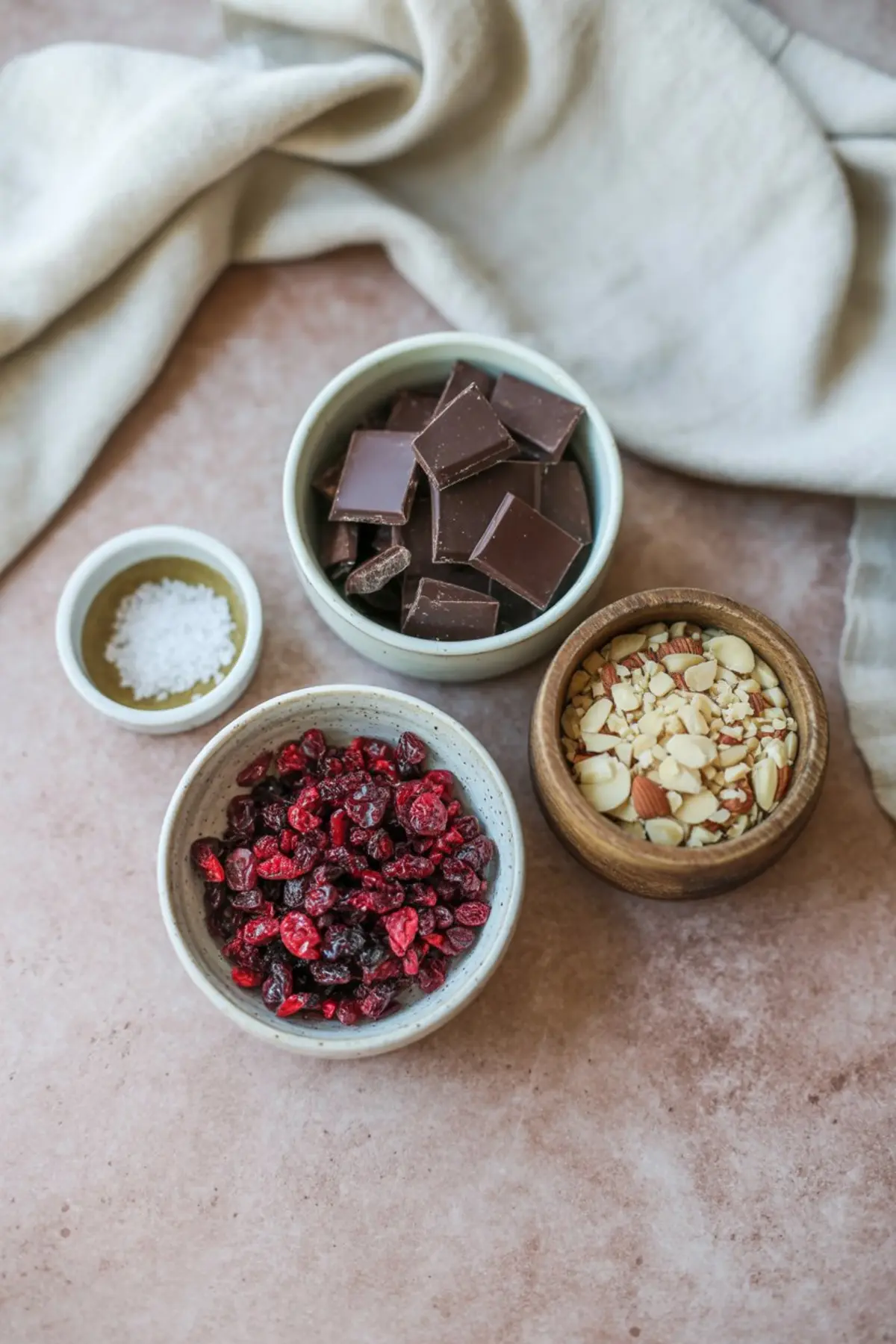 Overhead view of chocolate bark ingredients including dark chocolate chunks, dried cranberries, sliced almonds, and sea salt in small bowls on a pink background.
