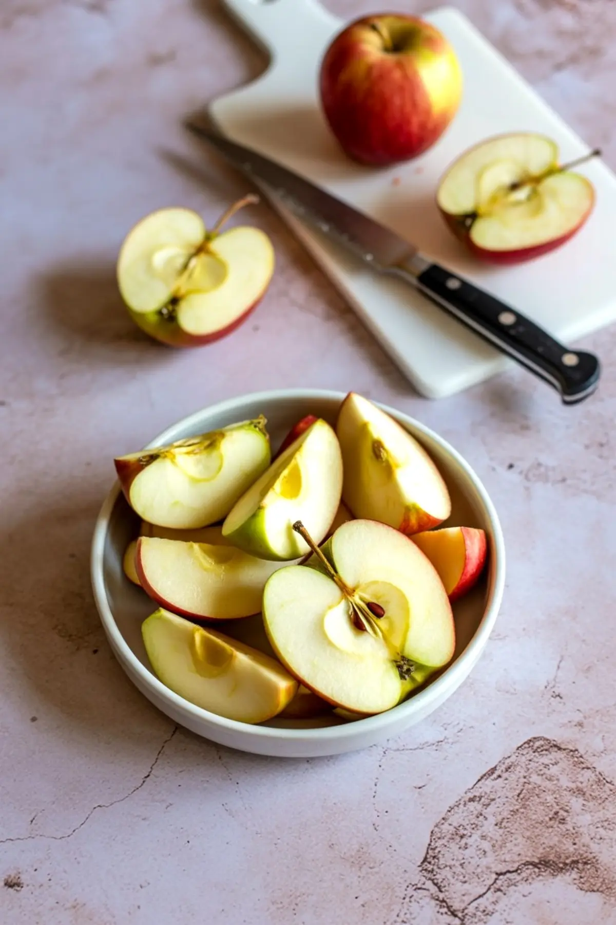 Sliced red and green apples in a white bowl with a cutting board, whole apple, and knife in the background on a light textured surface.