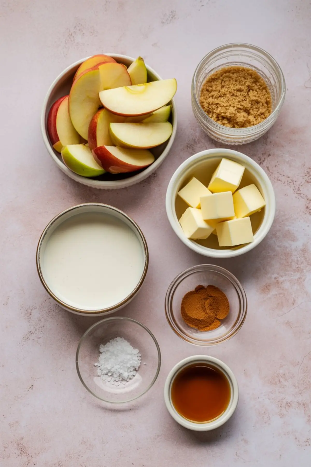 Flat lay of ingredients for cinnamon apples, including sliced red and green apples, brown sugar, cubed butter, heavy cream, ground cinnamon, vanilla extract, and cornstarch, all arranged on a pink background.