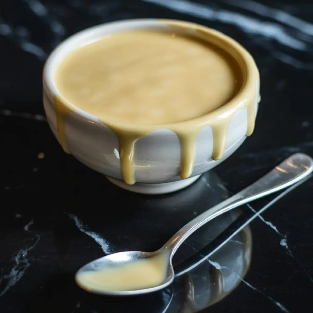 Bowl of creamy vanilla glaze or icing dripping slightly over the rim, placed on a glossy black marble countertop beside a silver spoon with a small scoop of glaze.