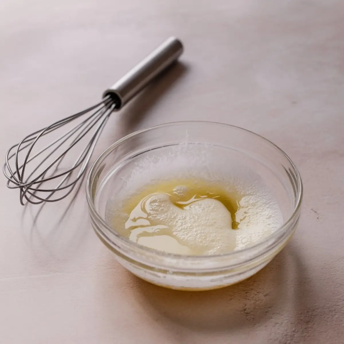 A small glass bowl with lightly frothed egg white mixture beside a stainless steel whisk, set on a soft blush background.