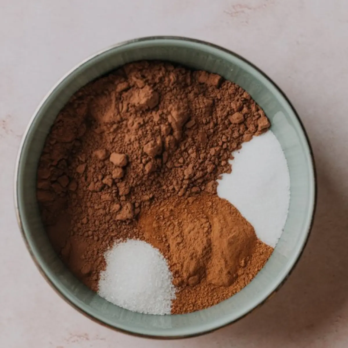 A green ceramic bowl filled with unsweetened cocoa powder, white sugar, and ground cinnamon arranged in separate sections.