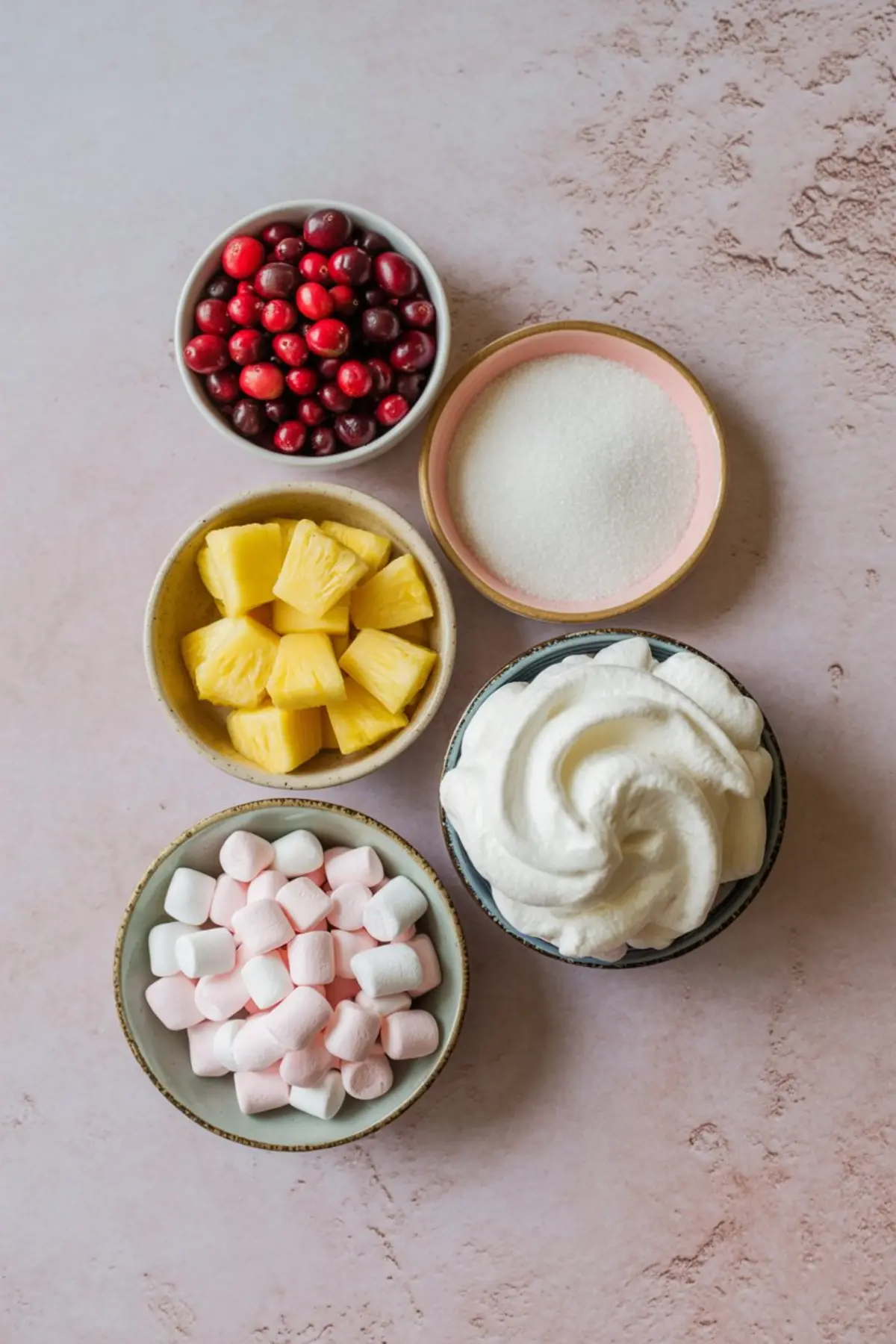 Five small bowls arranged on a pink surface containing cranberries, granulated sugar, chopped pineapple, whipped topping, and pastel mini marshmallows.
