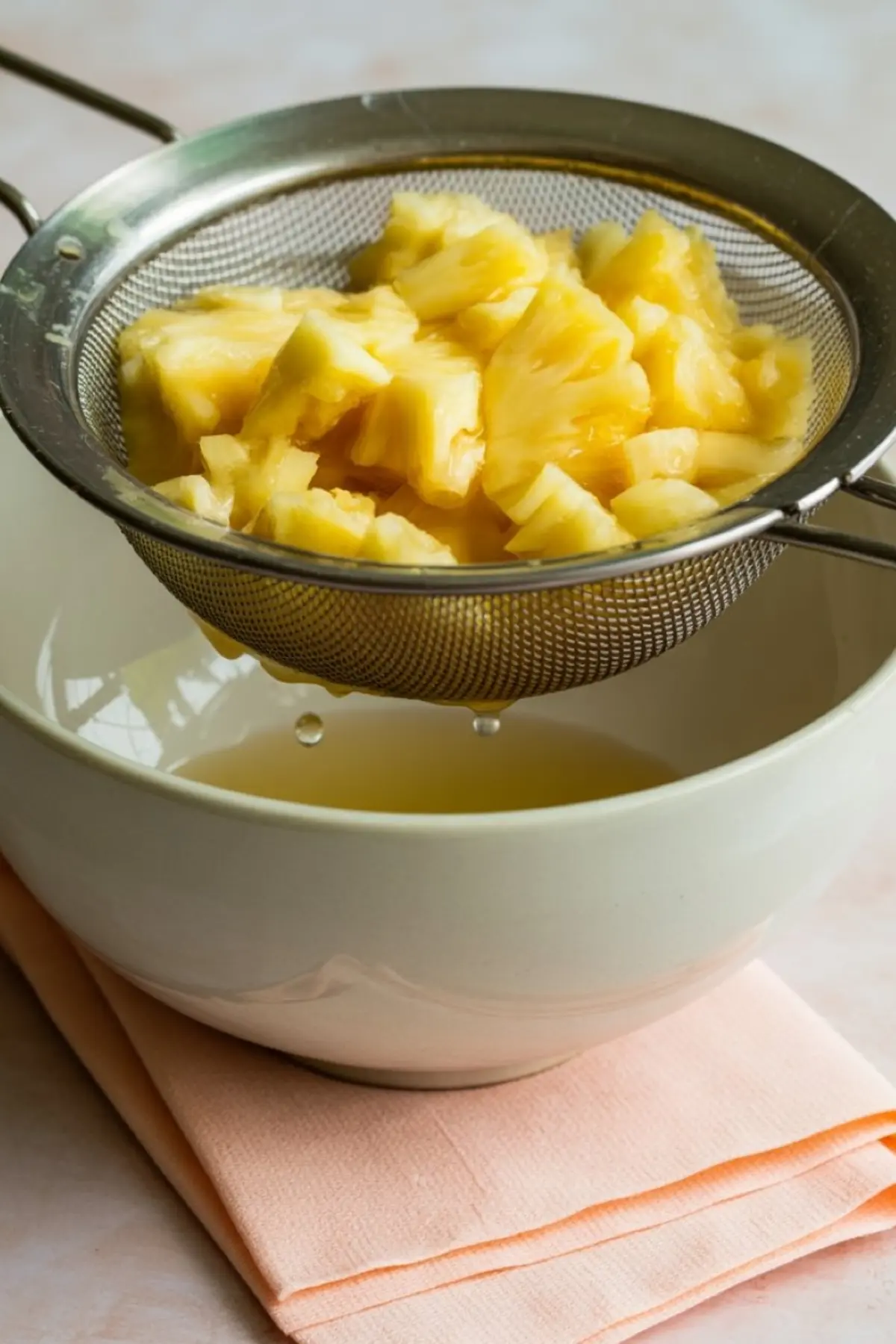 Chunks of fresh pineapple draining in a fine mesh strainer set over a ceramic bowl, with juice dripping into the bowl below on a peach cloth napkin.

