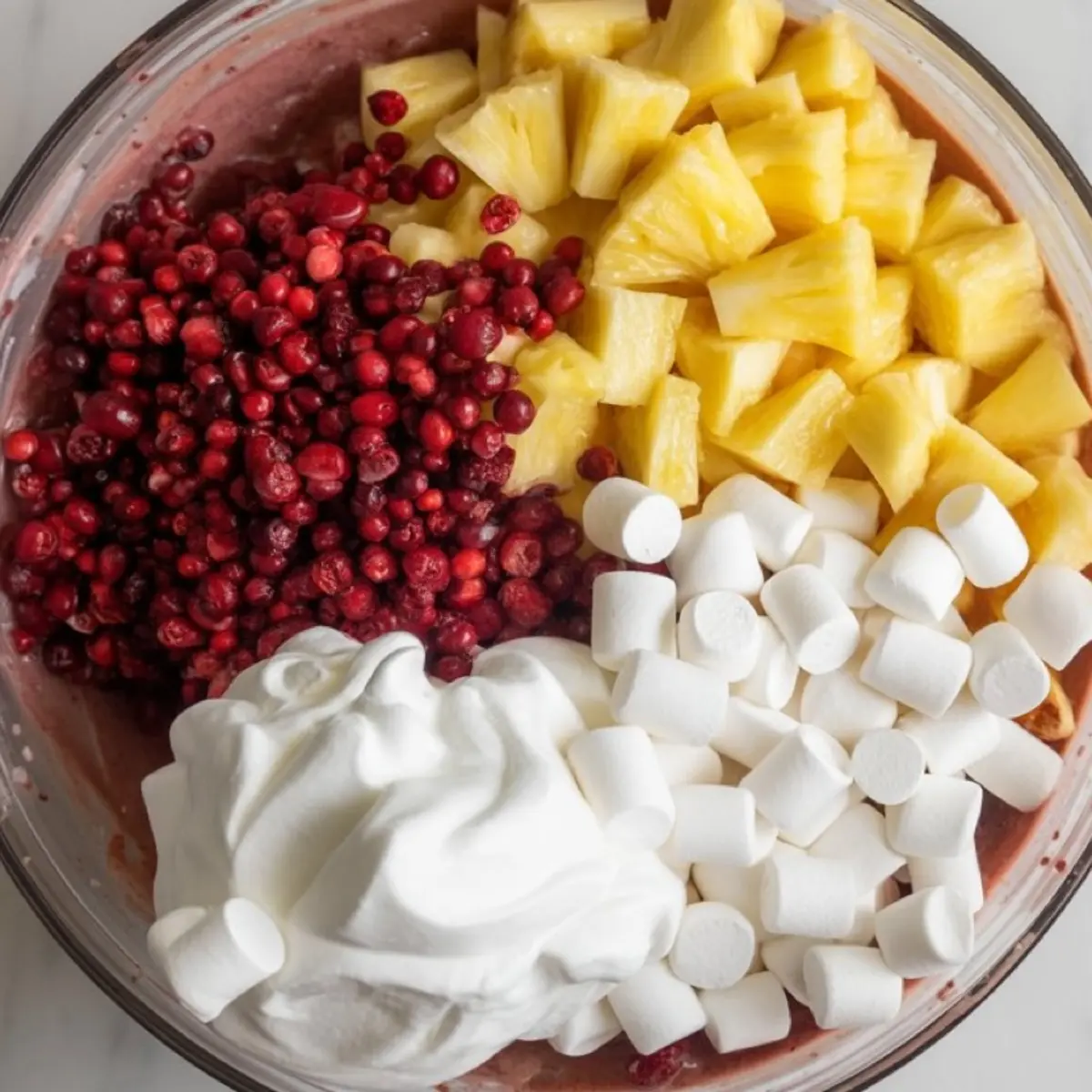 Glass mixing bowl filled with ingredients for cranberry fluff salad including chopped pineapple, whole cranberries, mini marshmallows, and whipped topping.
