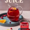 Glass pitcher and drinking glass filled with cranberry juice, garnished with fresh cranberries and orange slices, styled on a blue tray with scattered cranberries and citrus wedges, with the text “Cranberry Juice.”