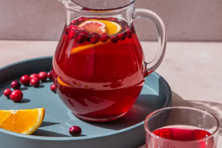 Glass pitcher and drinking glass filled with cranberry juice, garnished with fresh cranberries and orange slices, styled on a blue tray with scattered cranberries and citrus wedges, with the text “Cranberry Juice.”