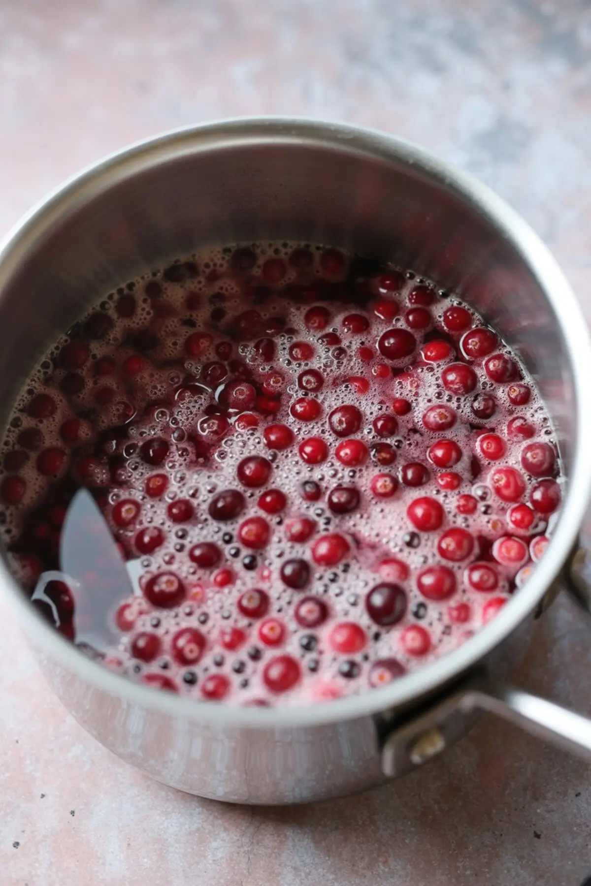 Cranberries simmering in a stainless steel saucepan with bubbling liquid on a kitchen counter.
