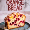 Moist cranberry orange bread slice on a blue plate, showing vibrant red cranberries and orange zest inside a soft, golden loaf, topped with citrus glaze and text above reading “Cranberry Orange Bread.”