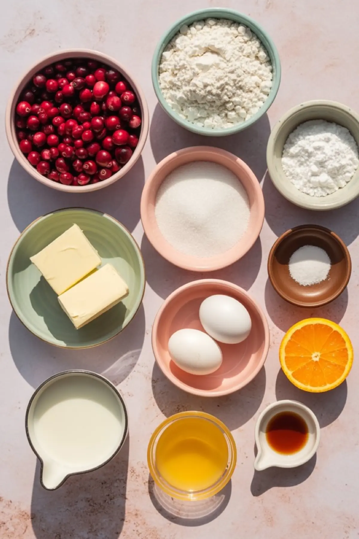 Top-down view of baking ingredients in colorful bowls including cranberries, flour, sugar, butter, eggs, orange juice, milk, vanilla extract, and a halved orange on a pastel background.
