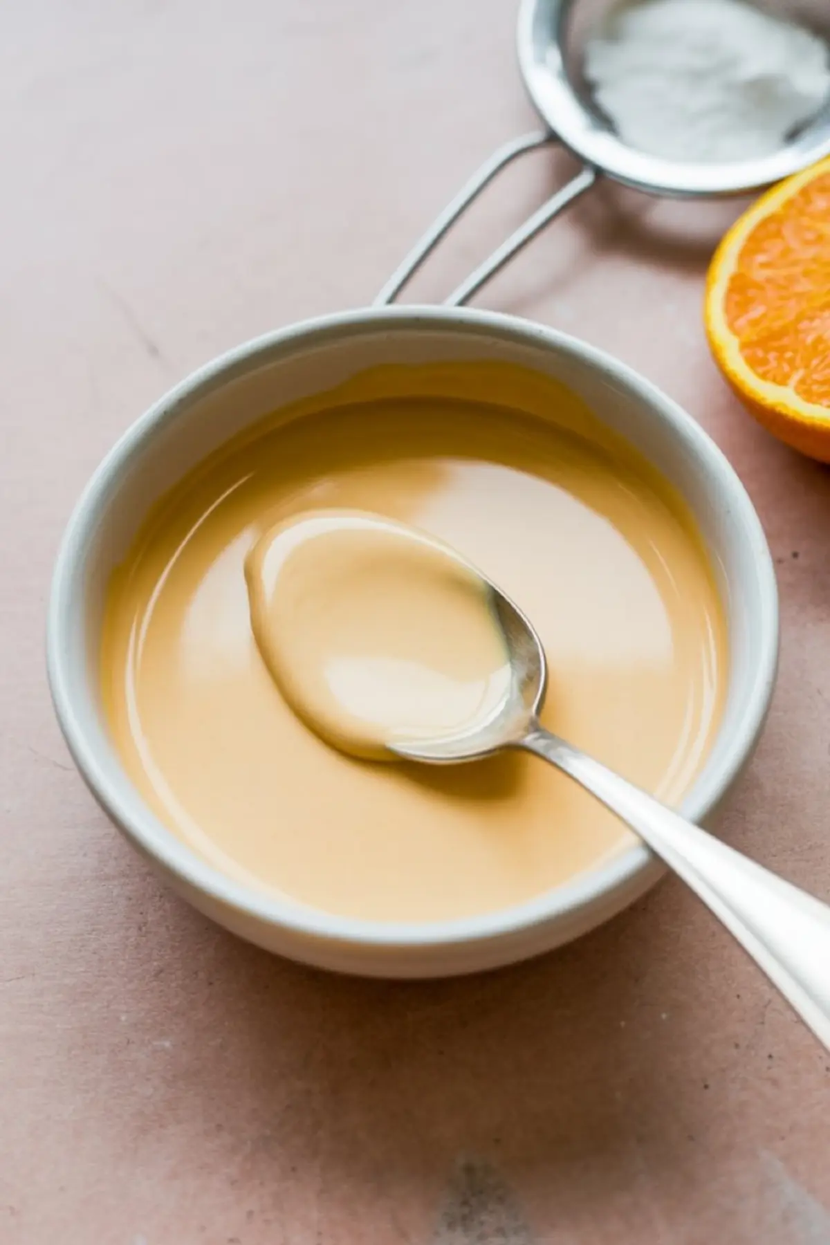 Close-up of creamy orange glaze in a white bowl with a spoon, set on a pink background with a halved orange and metal strainer nearby.
