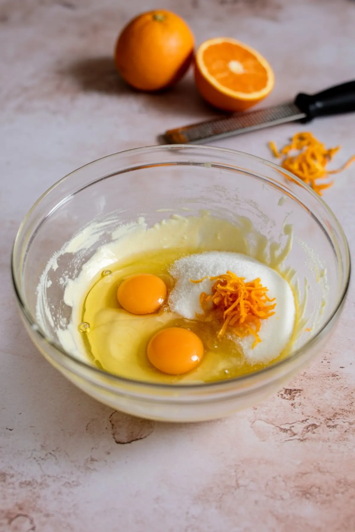 Glass mixing bowl with eggs, sugar, orange zest, and wet ingredients for baking, surrounded by a halved orange, zester, and orange peel on a marble surface.
