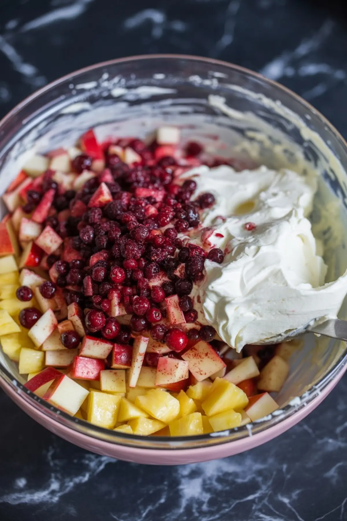 Large mixing bowl with diced apples, pineapple chunks, cranberries, and a dollop of whipped cream on a dark marble surface.