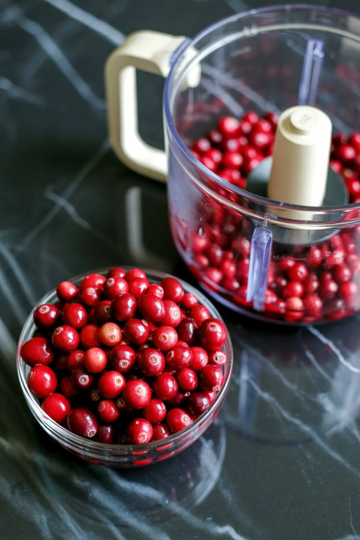 Fresh cranberries in a small glass bowl next to a food processor partially filled with cranberries on a black marble countertop.