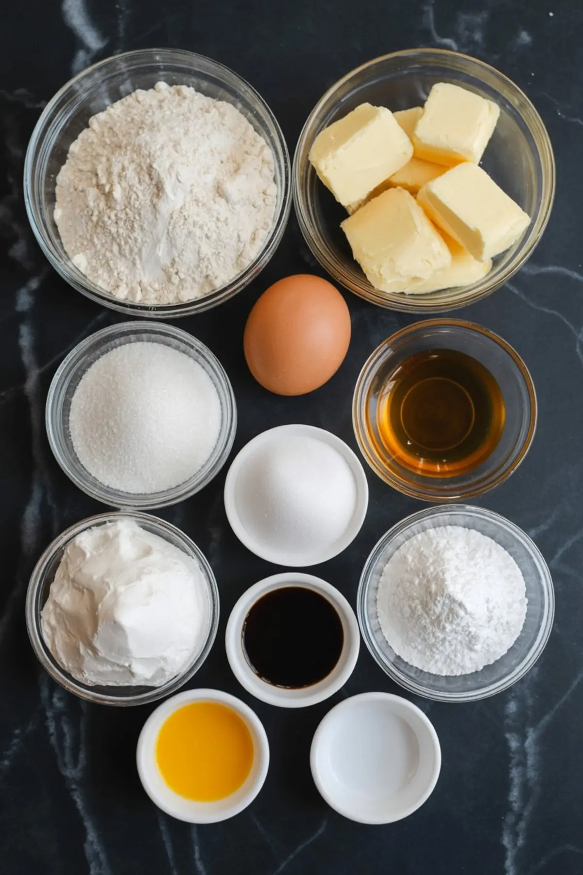 Overhead view of sugar cookie ingredients including flour, butter, sugar, egg, vanilla extract, powdered sugar, sour cream, and baking essentials arranged in glass bowls.