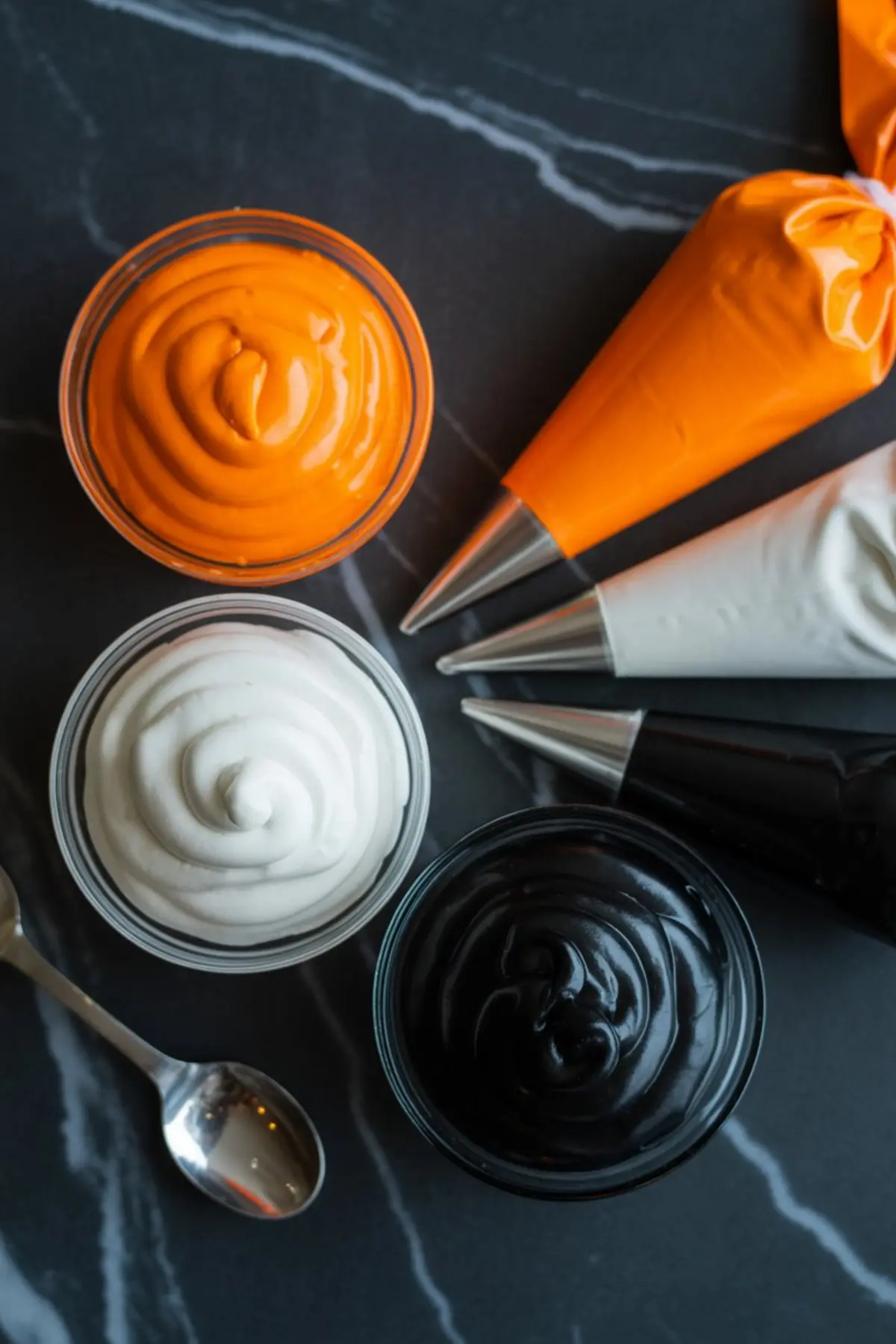 Bowls of vibrant cookie icing in orange, white, and black with matching piping bags ready for decorating Halloween cookies.