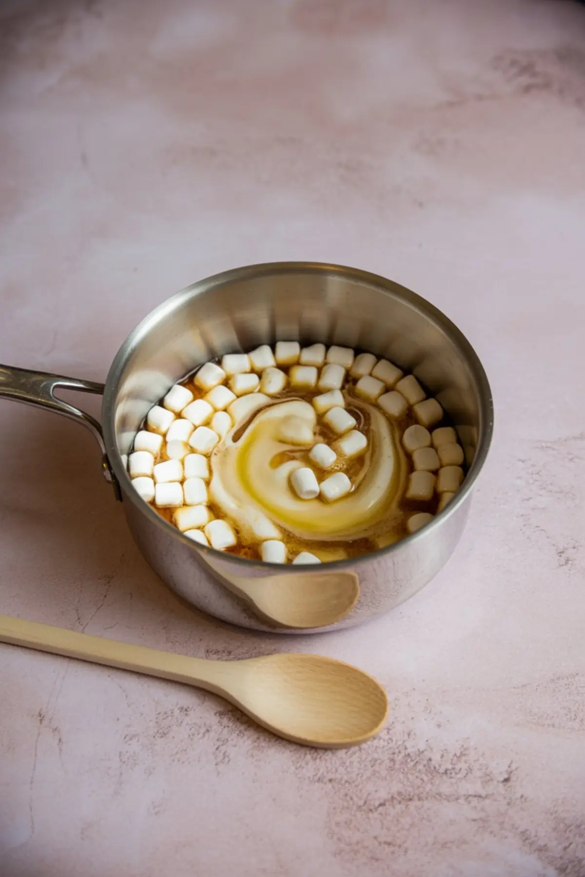 Mini marshmallows melting with butter in a silver saucepan, placed next to a wooden spoon on a pink countertop.