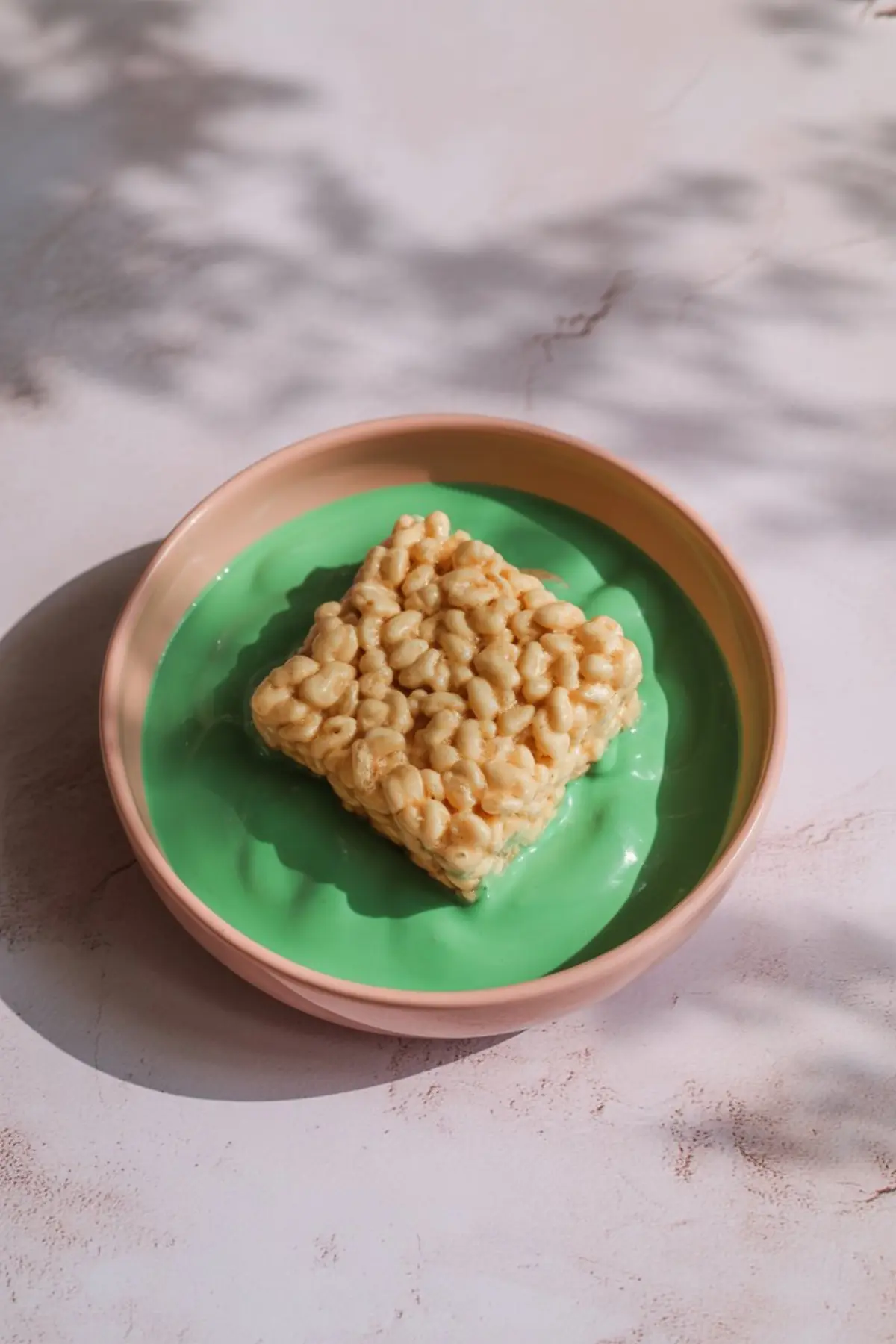 Square Rice Krispie treat dipped in melted green candy coating in a shallow pink bowl, shot in natural daylight.