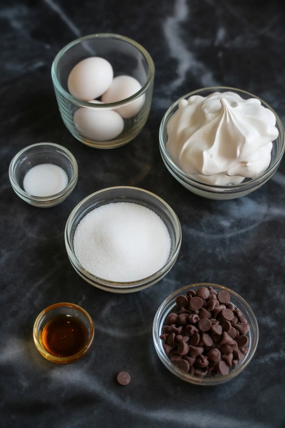 Flat lay of ghost meringue ingredients including eggs, granulated sugar, vanilla extract, chocolate chips, cream of tartar, and whipped meringue in glass bowls.