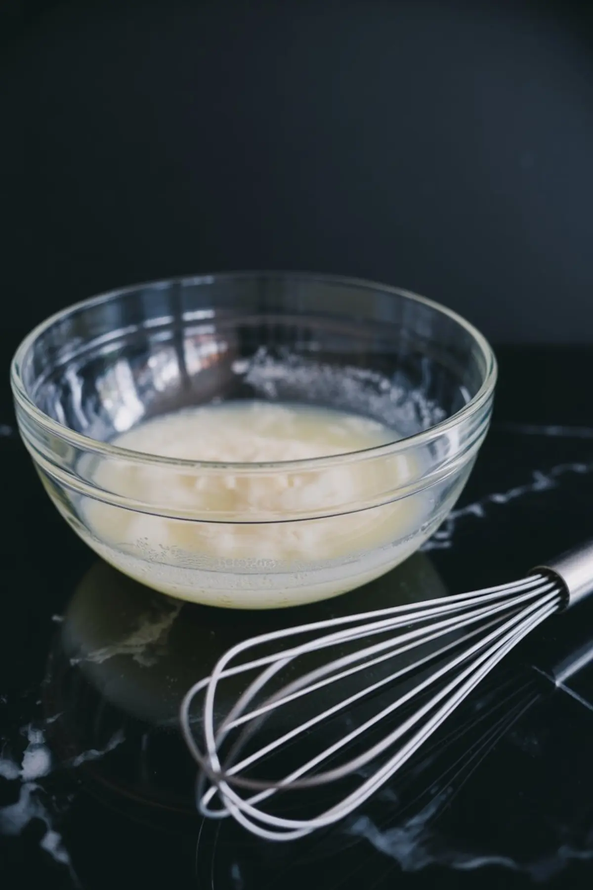 Unwhipped egg whites and sugar mixture in a clear glass bowl with a metal whisk nearby, set on a black countertop for meringue preparation.