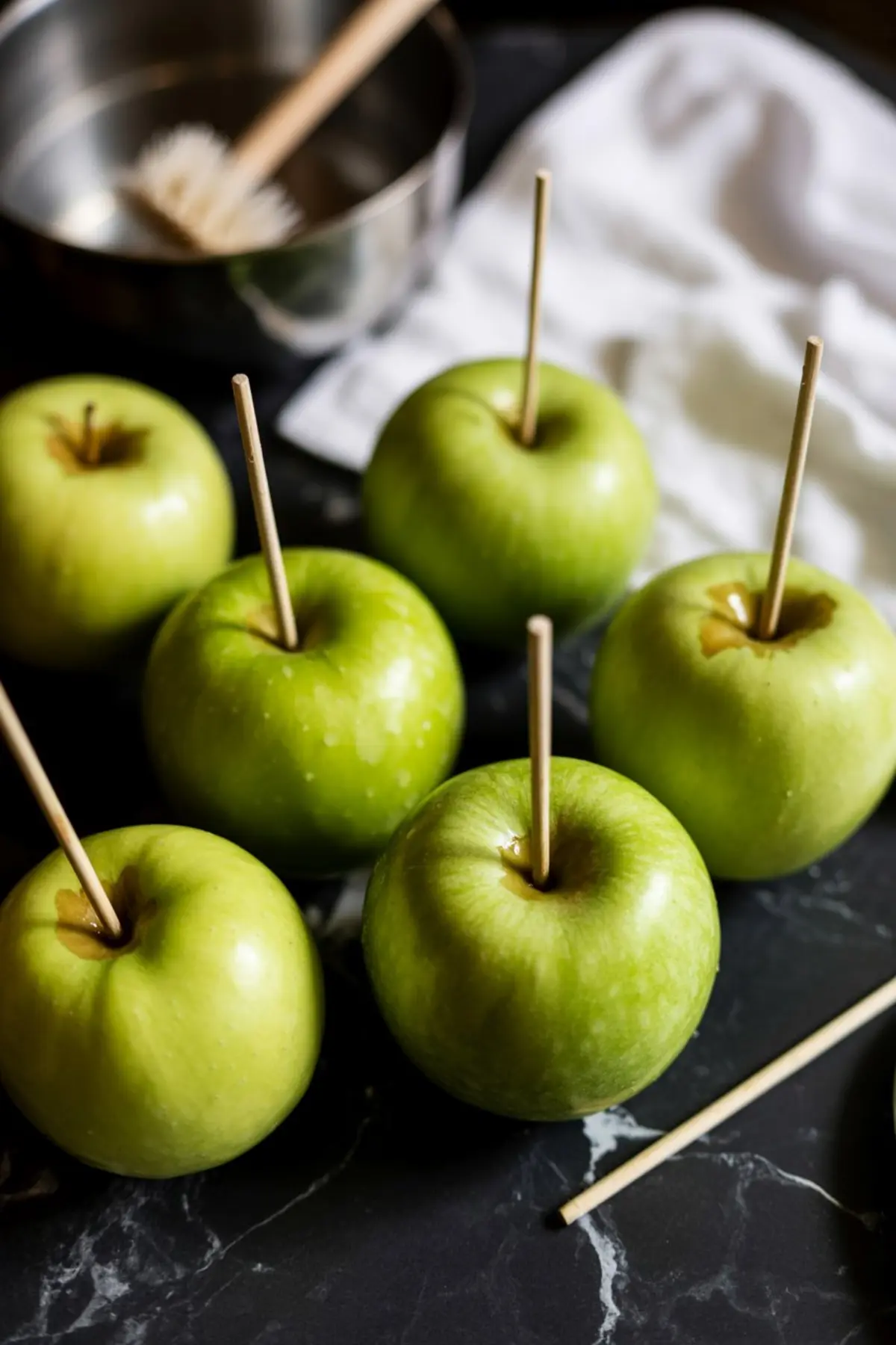 Fresh Granny Smith apples stand upright on black marble after skewering with wooden sticks, waiting for caramel coating.