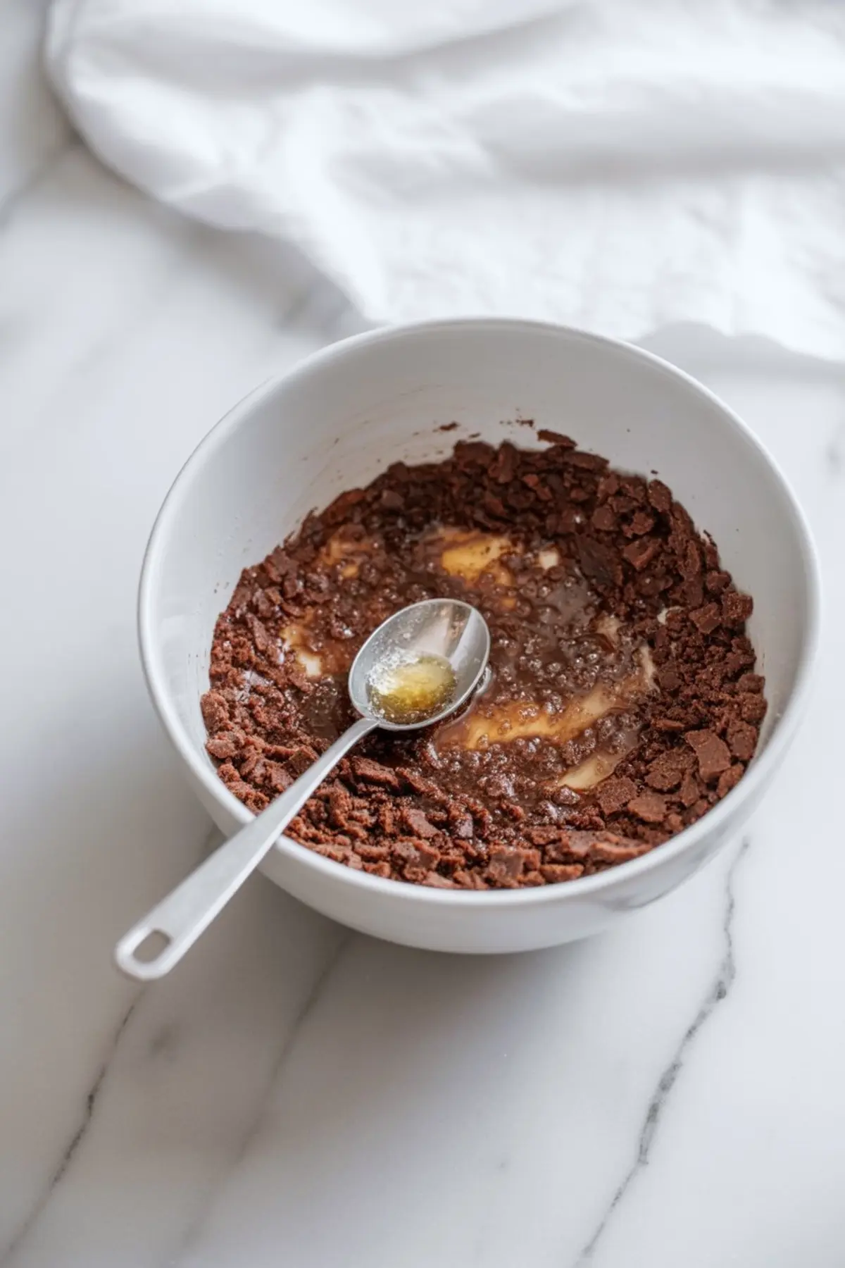 Partially mixed chocolate cookie crumbs and butter in a white bowl with a spoon, prepared for a no-bake cheesecake base.
