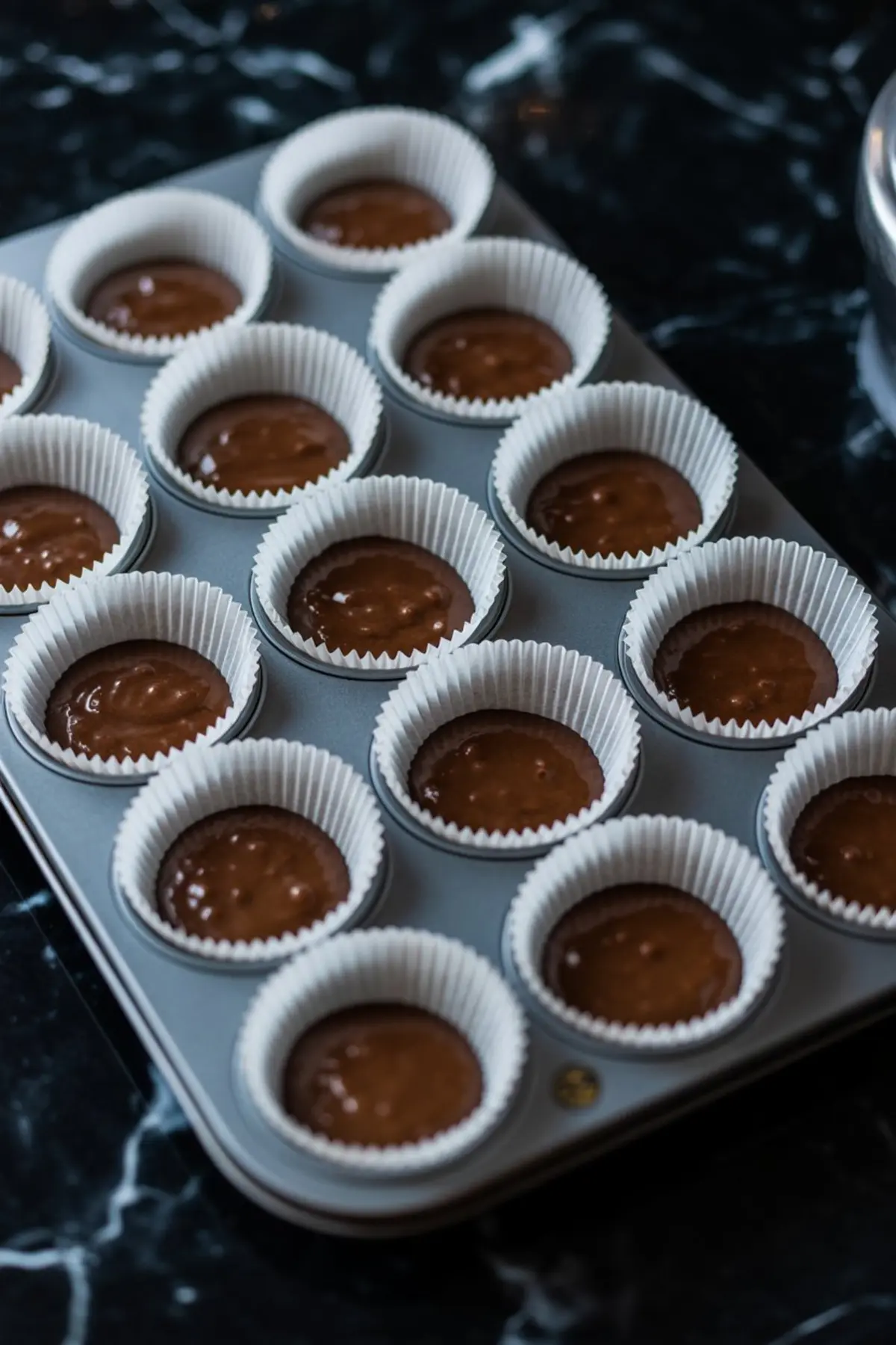 A nonstick muffin tray lined with white paper cupcake liners is filled with smooth chocolate batter, arranged neatly in rows, ready to go into the oven for baking cupcakes.