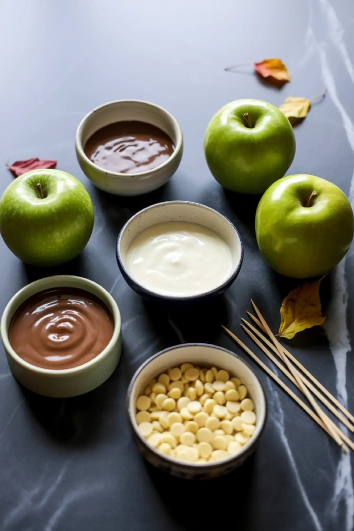 Flat lay of caramel apple dipping setup featuring fresh green apples, bowls of melted milk chocolate, white chocolate, dark chocolate, and white chocolate chips on a black marble surface with wooden skewers and autumn leaves.
