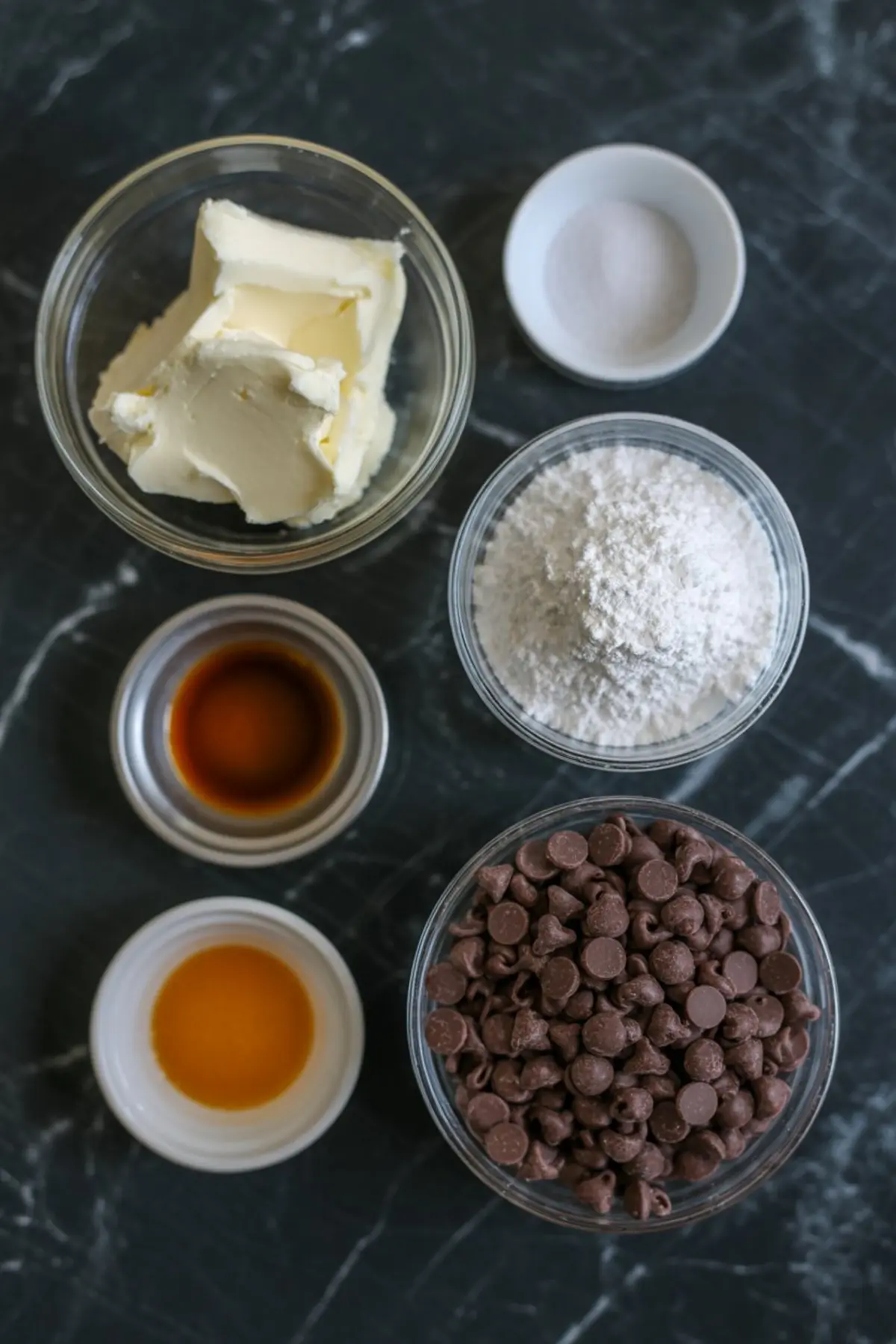 Flat lay of baking ingredients on a dark countertop, including butter, vanilla extract, salt, flour, chocolate chips, and caramel sauce, used for creating a sweet dessert dip or cookie dough treat.