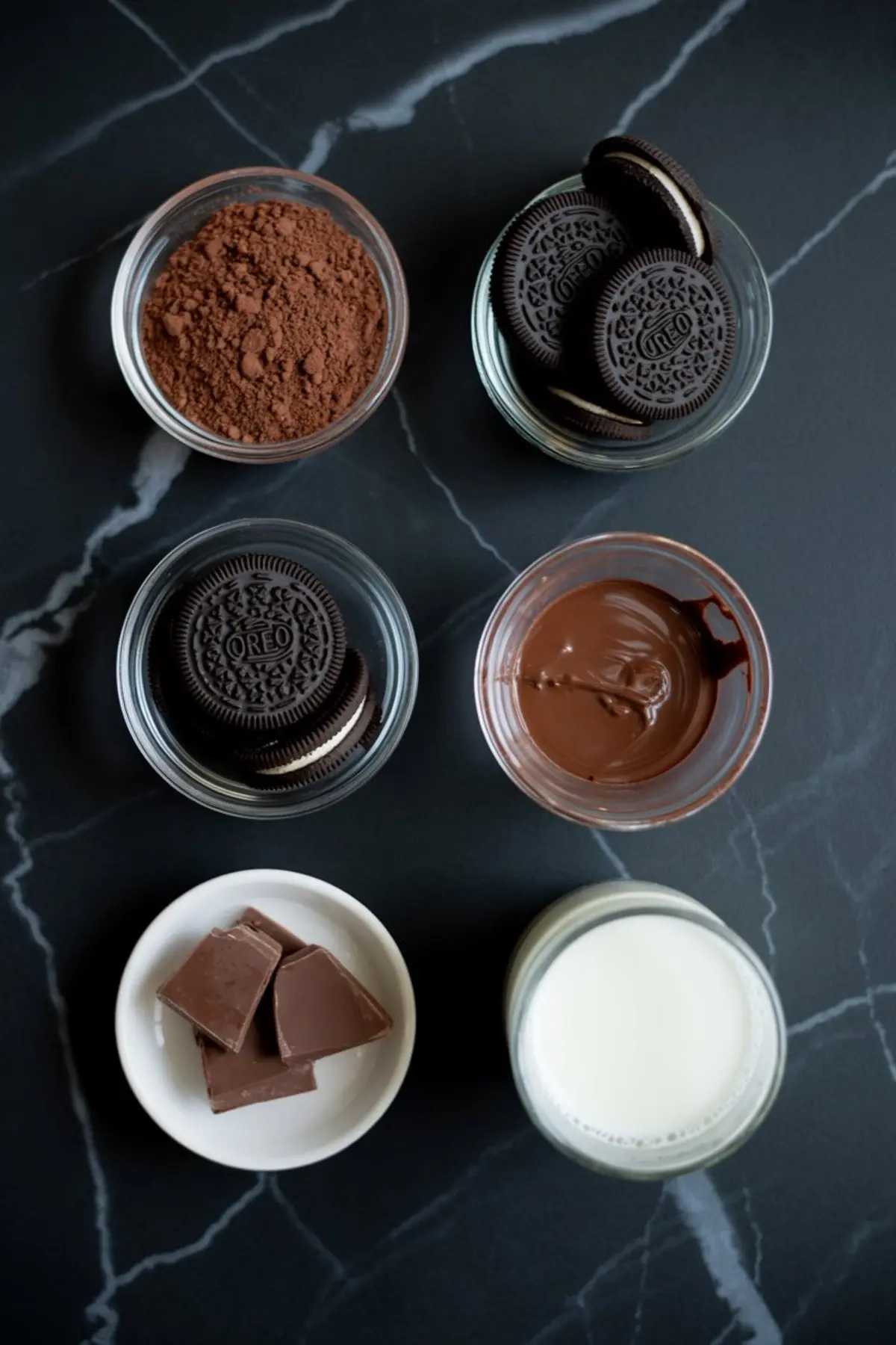 Flat lay of Halloween dirt cup ingredients including Oreo cookies, cocoa powder, melted chocolate, milk chocolate chunks, and a glass of milk arranged in bowls on black marble surface.