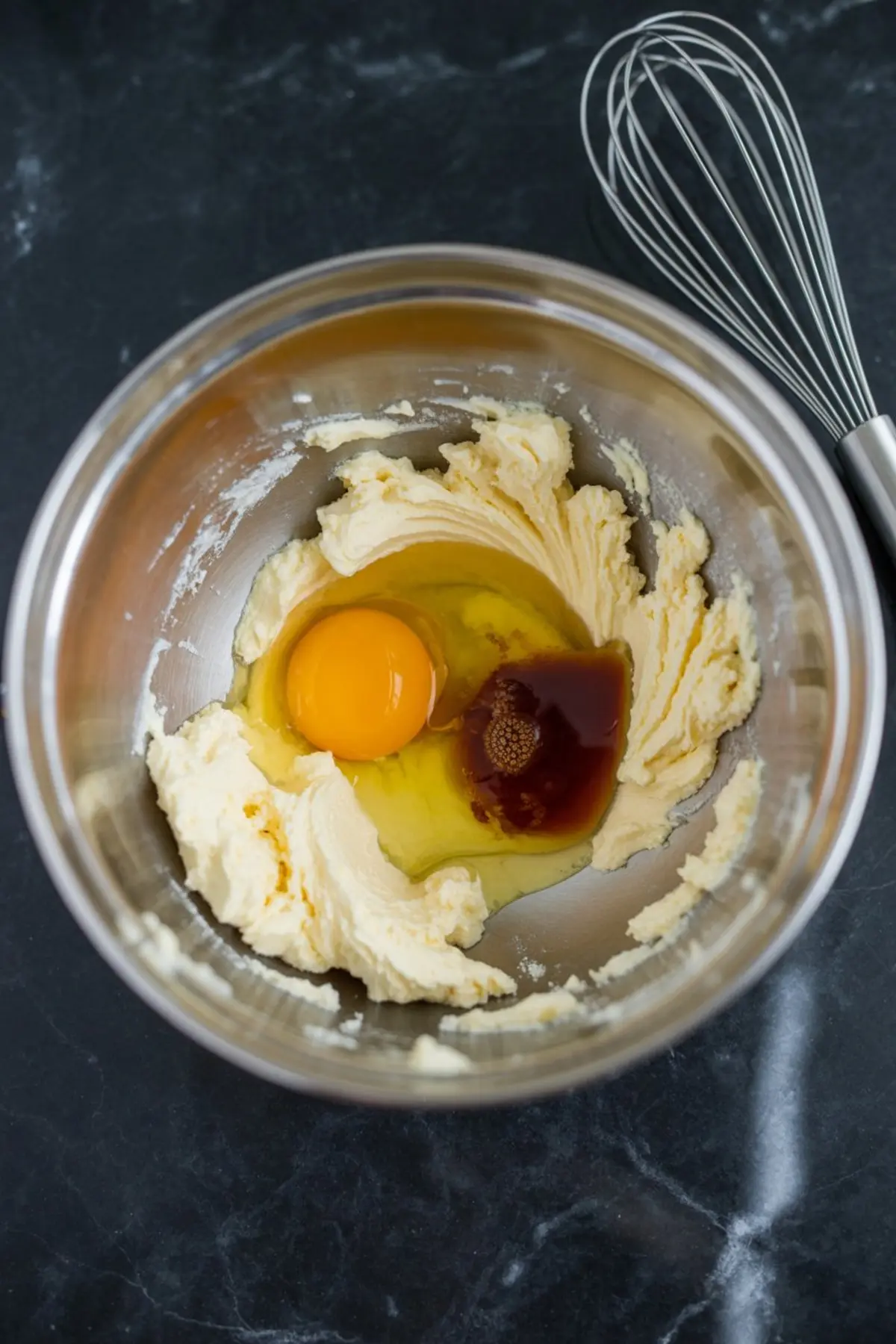 Stainless steel mixing bowl containing creamed butter and sugar, with a cracked egg and vanilla extract added, set beside a metal whisk on a dark surface.