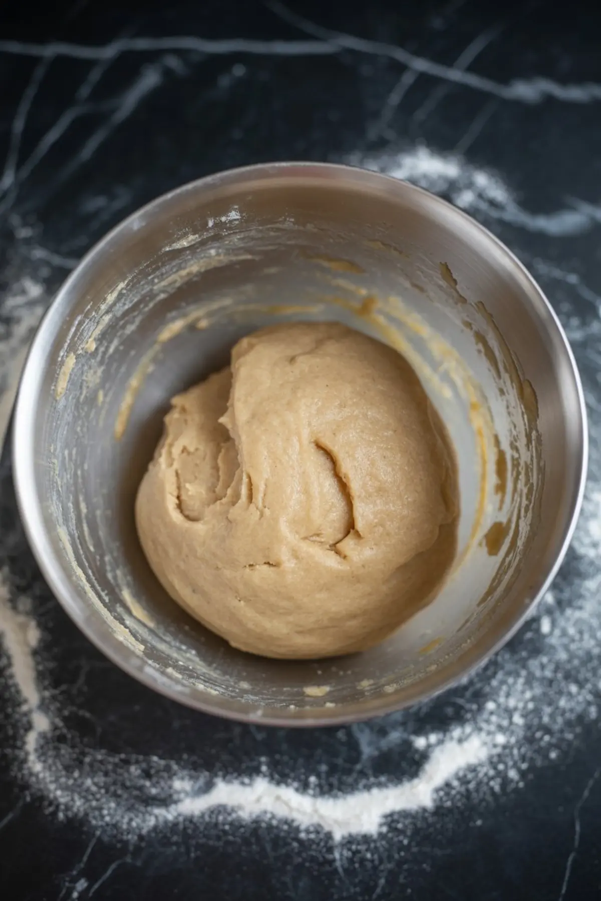 Smooth cookie dough formed into a ball resting in a metal bowl, with flour dusted around the dark countertop background.