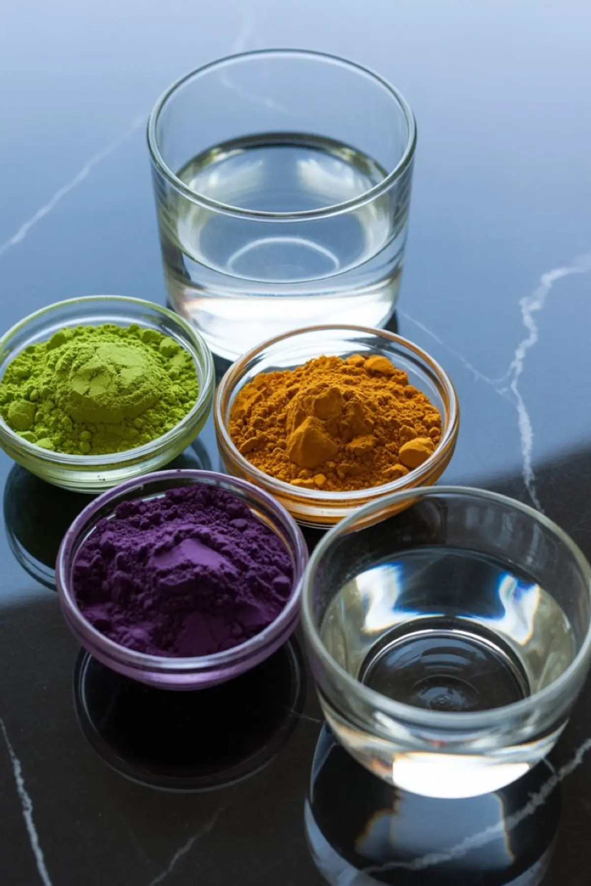 Group of small glass bowls filled with colorful powders in green, orange, and purple, arranged alongside three glasses of clear liquid on a glossy black surface.