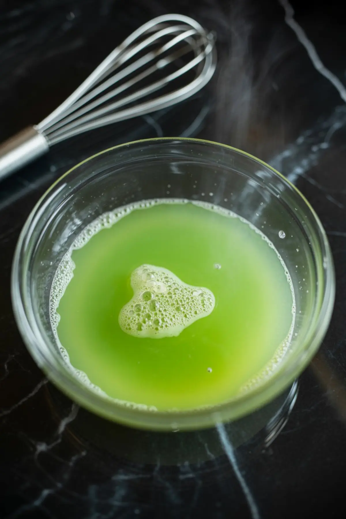 Transparent bowl of bright green liquid with foam bubbles floating on top, set on a black marble surface beside a stainless steel whisk.