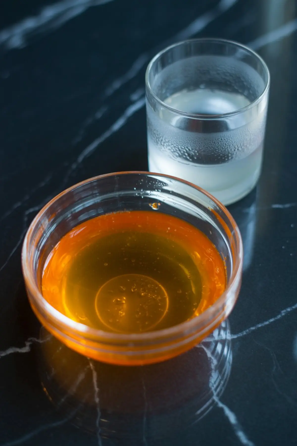 Glass bowl with golden amber liquid placed next to a clear glass of translucent liquid, both resting on a dark marble countertop.