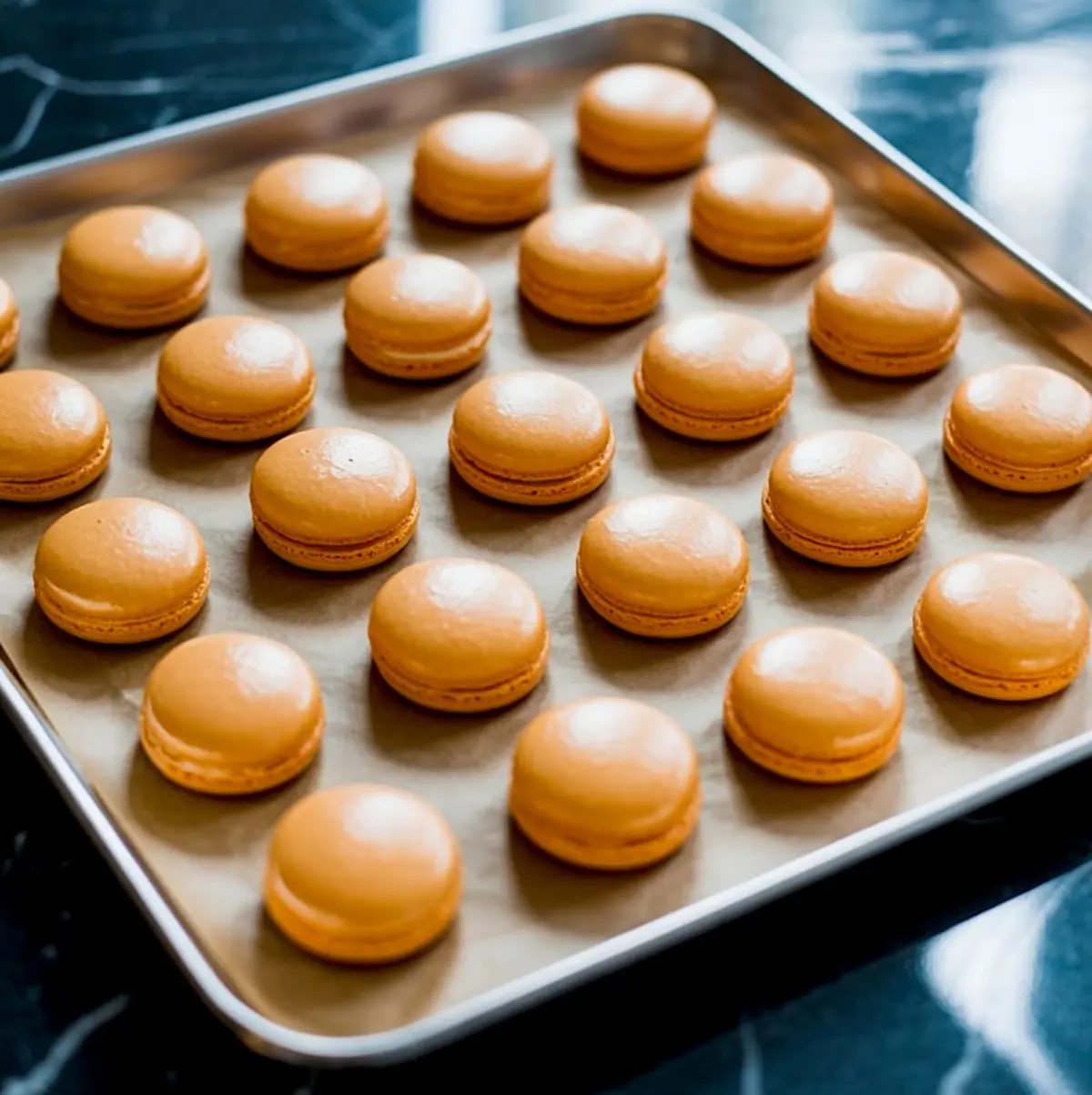 Parchment-lined baking sheet displays evenly spaced orange macaron shells cooling, smooth tops awaiting pumpkin face decoration.