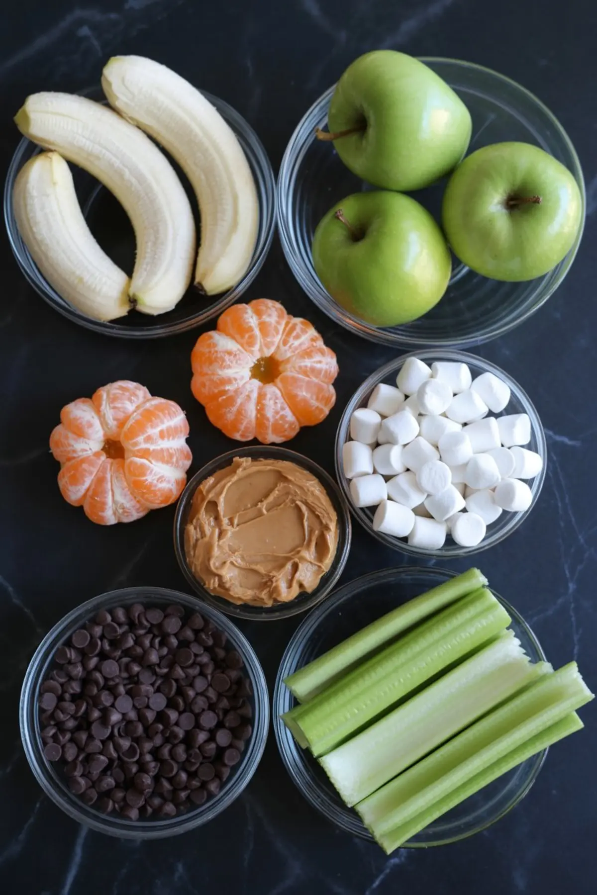 Flat lay of healthy snack ingredients including bananas, green apples, peeled mandarins, mini marshmallows, peanut butter, celery sticks, and chocolate chips in clear glass bowls on a dark countertop.