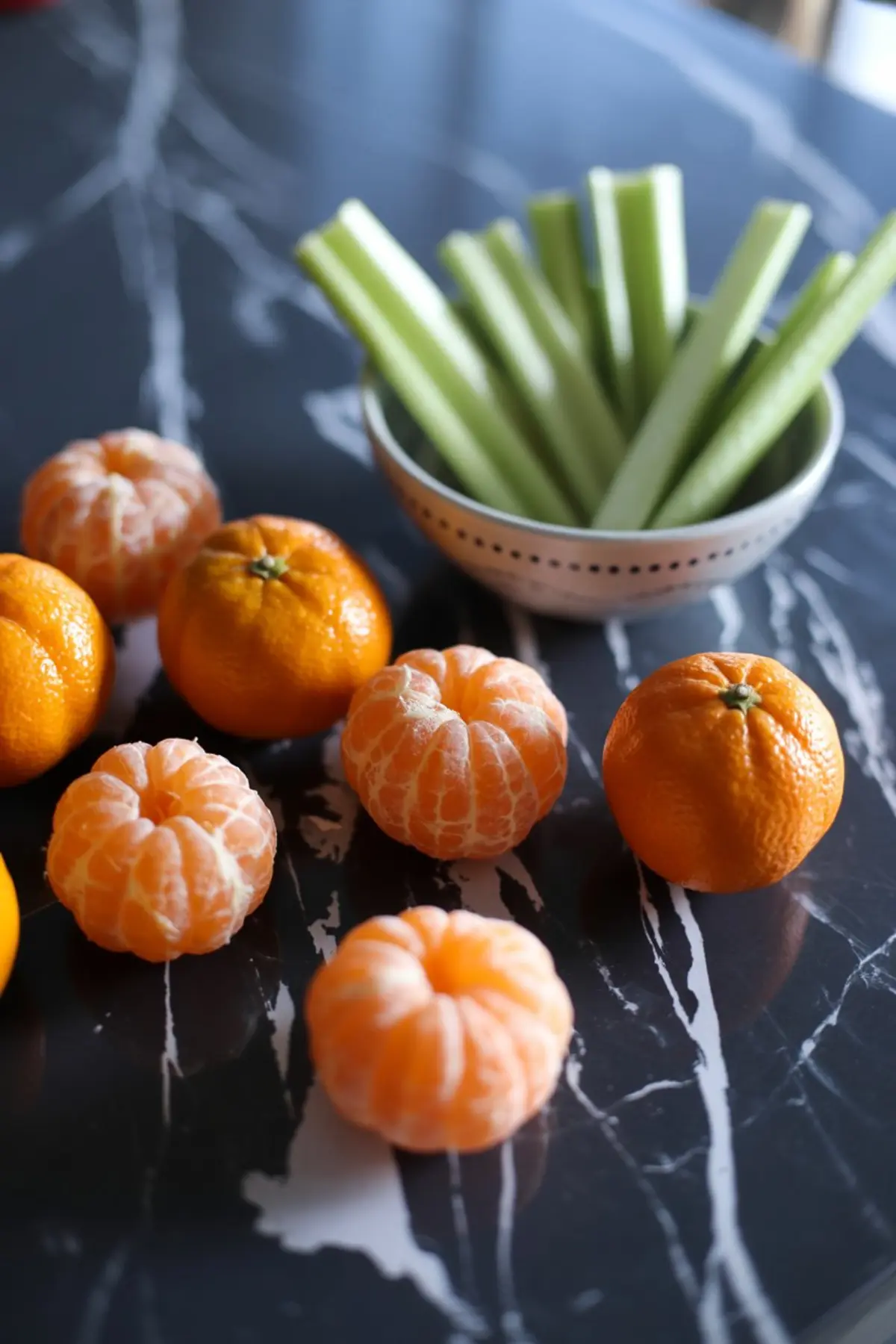 Fresh whole and peeled mandarins arranged on a black marble counter beside a white bowl filled with crisp celery sticks.