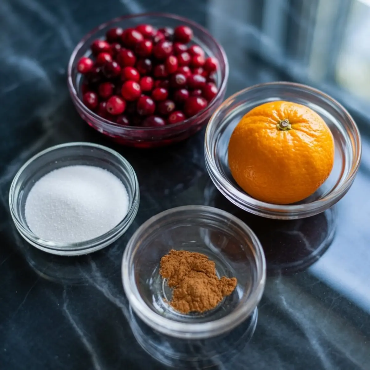 Glass bowls on a dark marble surface hold fresh cranberries, a whole orange, granulated sugar, and ground cinnamon, showcasing ingredients for homemade cranberry sauce.