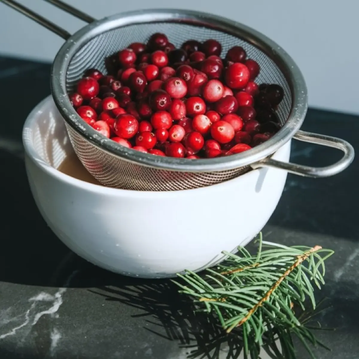 A stainless steel strainer filled with rinsed cranberries rests over a white bowl, with a pine sprig beside it on a dark marble countertop.